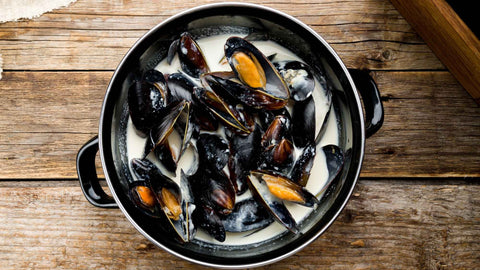 (Cocotte en fonte : moules brillantes ouvertes, sauce crème-cidre perlée, brins de persil, quartier de citron, panier de frites à côté.) 📷